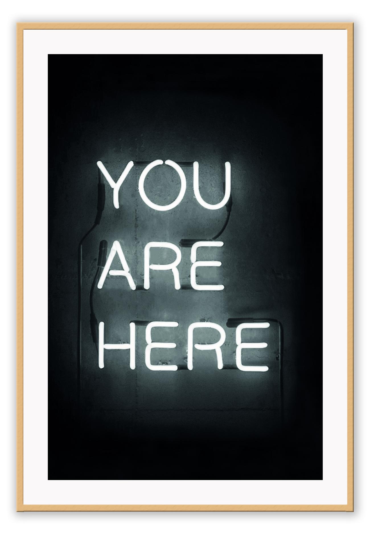 A black and white, scandi typography wall art with YOU ARE HERE neon letter lighting installation on a black wall background. 